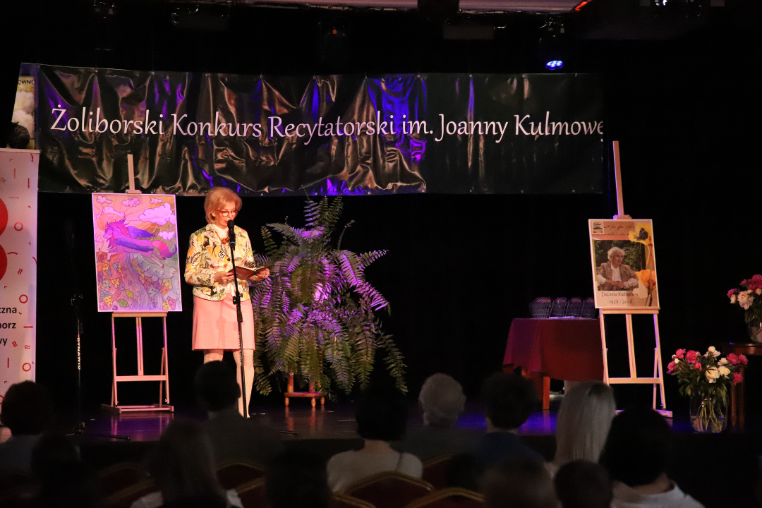 Zdjęcie z III Żoliborskiego Konkursu Recytatorskiego im. Joanny Kulmowej zorganizowanego przez Bibliotekę Publiczną w Dzielnicy Żoliborz, Społeczny Dom Kultury WSM oraz Fundację Joanny i Jana Kulmów. Kobieta na scenie mówi do mikrofonu.