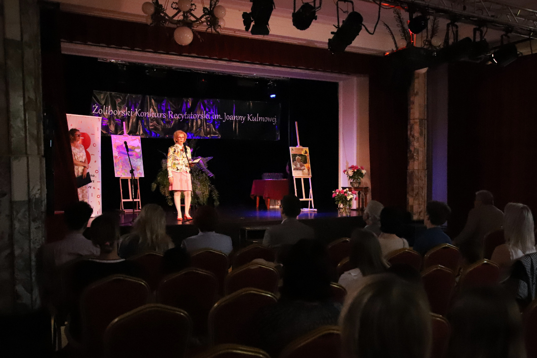 Zdjęcie z III Żoliborskiego Konkursu Recytatorskiego im. Joanny Kulmowej zorganizowanego przez Bibliotekę Publiczną w Dzielnicy Żoliborz, Społeczny Dom Kultury WSM oraz Fundację Joanny i Jana Kulmów. Kobieta na scenie mówi do mikrofonu.