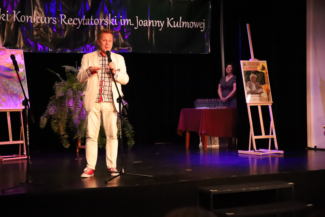 Zdjęcie z III Żoliborskiego Konkursu Recytatorskiego im. Joanny Kulmowej zorganizowanego przez Bibliotekę Publiczną w Dzielnicy Żoliborz, Społeczny Dom Kultury WSM oraz Fundację Joanny i Jana Kulmów. Mężczyzna na scenie mówi do mikrofonu.