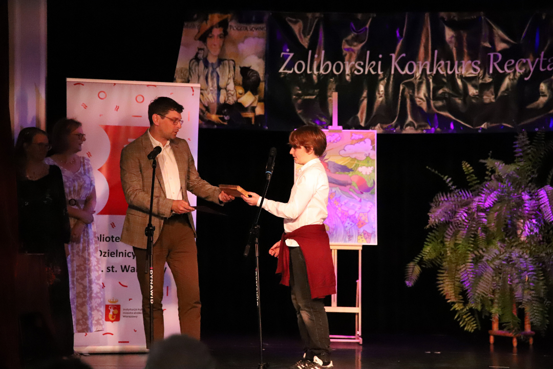 Zdjęcie z III Żoliborskiego Konkursu Recytatorskiego im. Joanny Kulmowej zorganizowanego przez Bibliotekę Publiczną w Dzielnicy Żoliborz, Społeczny Dom Kultury WSM oraz Fundację Joanny i Jana Kulmów. Mężczyzna na scenie podaje drewnianą deskę chłopakowi.