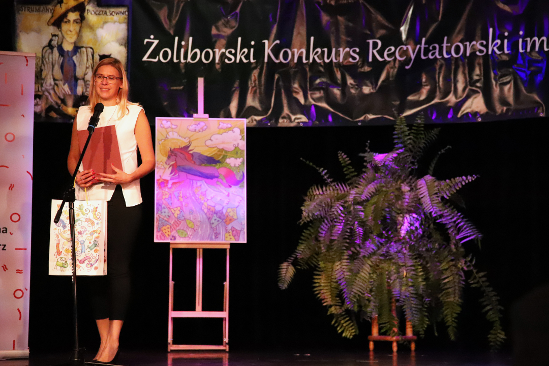 Zdjęcie z III Żoliborskiego Konkursu Recytatorskiego im. Joanny Kulmowej zorganizowanego przez Bibliotekę Publiczną w Dzielnicy Żoliborz, Społeczny Dom Kultury WSM oraz Fundację Joanny i Jana Kulmów. Kobieta stoi na scenie i mówi do mikrofonu.