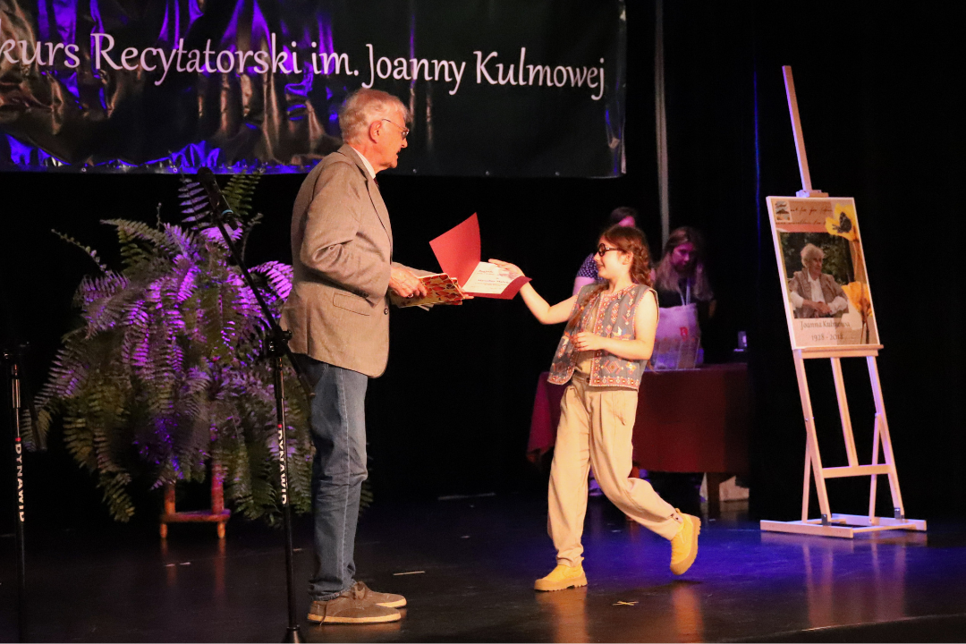 Zdjęcie z III Żoliborskiego Konkursu Recytatorskiego im. Joanny Kulmowej zorganizowanego przez Bibliotekę Publiczną w Dzielnicy Żoliborz, Społeczny Dom Kultury WSM oraz Fundację Joanny i Jana Kulmów. Mężczyzna na scenie podaje drewnianą deskę chłopakowi.