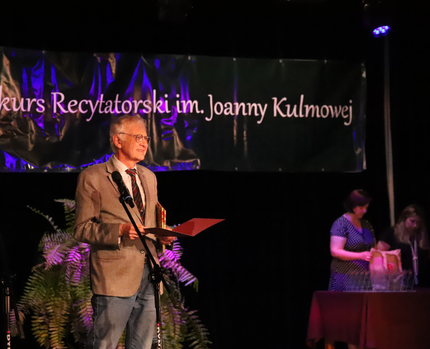Zdjęcie z III Żoliborskiego Konkursu Recytatorskiego im. Joanny Kulmowej zorganizowanego przez Bibliotekę Publiczną w Dzielnicy Żoliborz, Społeczny Dom Kultury WSM oraz Fundację Joanny i Jana Kulmów. Mężczyzna na scenie mówi do mikrofonu.