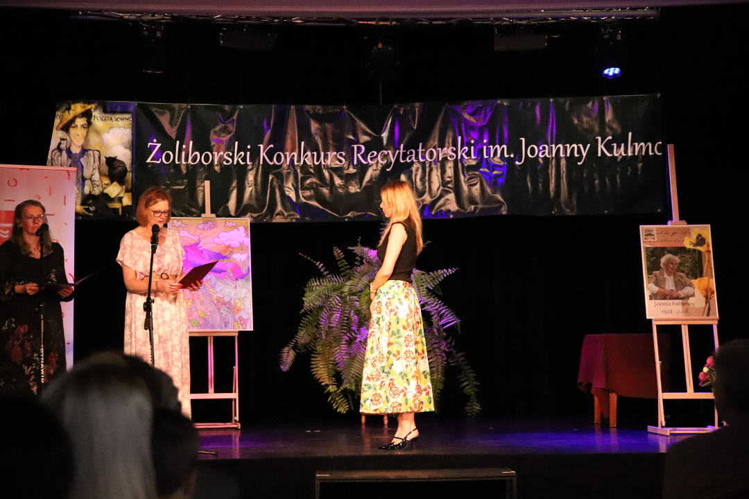 Zdjęcie z III Żoliborskiego Konkursu Recytatorskiego im. Joanny Kulmowej zorganizowanego przez Bibliotekę Publiczną w Dzielnicy Żoliborz, Społeczny Dom Kultury WSM oraz Fundację Joanny i Jana Kulmów. Trzy kobiety stoją na scenie i mówią do mikrofonu.