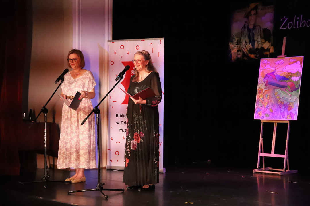 Zdjęcie z III Żoliborskiego Konkursu Recytatorskiego im. Joanny Kulmowej zorganizowanego przez Bibliotekę Publiczną w Dzielnicy Żoliborz, Społeczny Dom Kultury WSM oraz Fundację Joanny i Jana Kulmów. Dwie kobiety na scenie prezentują wydarzenie. Obok nich obraz z kolorowym koniem.