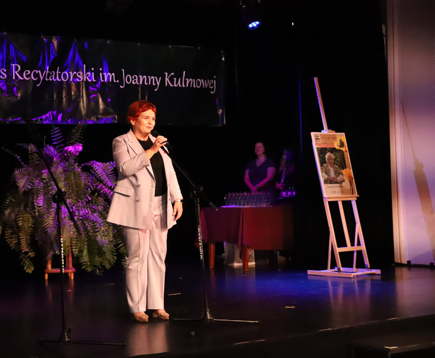 Zdjęcie z III Żoliborskiego Konkursu Recytatorskiego im. Joanny Kulmowej zorganizowanego przez Bibliotekę Publiczną w Dzielnicy Żoliborz, Społeczny Dom Kultury WSM oraz Fundację Joanny i Jana Kulmów. Kobieta na scenie mówi do mikrofonu.