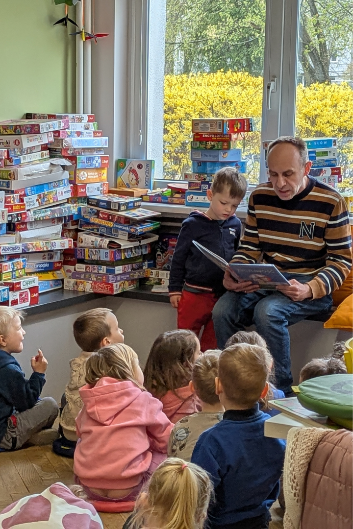 zdjęcie mężczyzny siedzącego i czytającego książkę siedzącym na podłodze dzieciom we wnętrzu biblioteki dla dzieci nr xv. w tle regały biblioteczne.