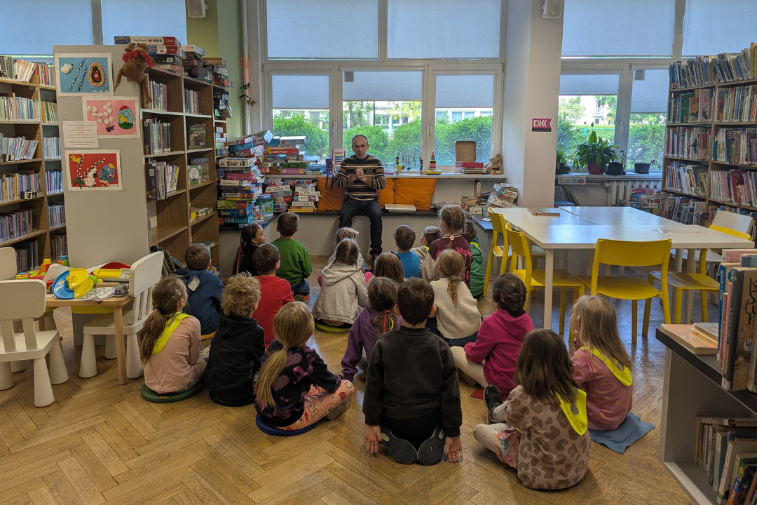 zdjęcie mężczyzny siedzącego i czytającego książkę siedzącym na podłodze dzieciom we wnętrzu biblioteki dla dzieci nr xv. w tle regały biblioteczne.