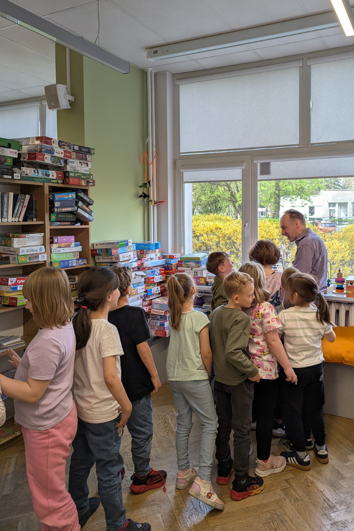 zdjęcie mężczyzny stojącego i czytającego książkę stojącym w kolejce na podłodze dzieciom we wnętrzu biblioteki dla dzieci nr xv. w tle regały biblioteczne.