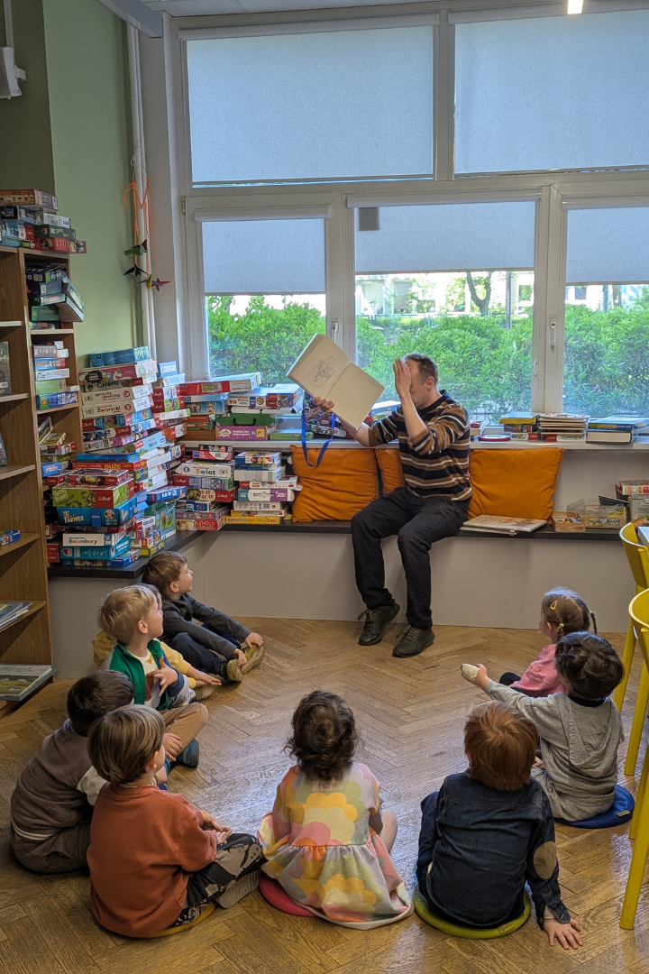 zdjęcie mężczyzny siedzącego i czytającego książkę siedzącym na podłodze dzieciom we wnętrzu biblioteki dla dzieci nr xv. w tle regały biblioteczne.