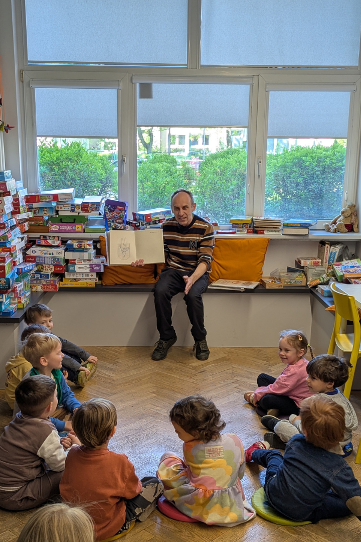 zdjęcie mężczyzny siedzącego i czytającego książkę siedzącym na podłodze dzieciom we wnętrzu biblioteki dla dzieci nr xv. w tle regały biblioteczne.