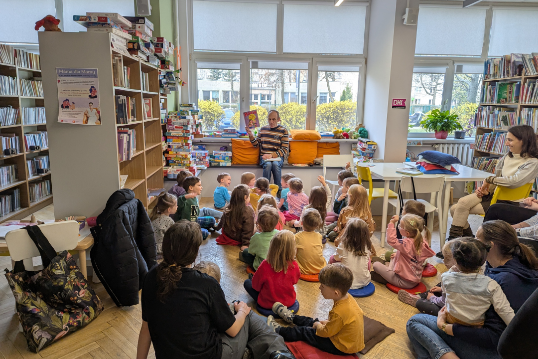 zdjęcie mężczyzny siedzącego i czytającego książkę siedzącym na podłodze dzieciom we wnętrzu biblioteki dla dzieci nr xv. w tle regały biblioteczne.