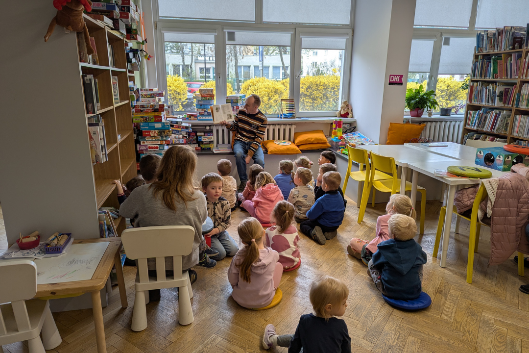 zdjęcie mężczyzny siedzącego i czytającego książkę siedzącym na podłodze dzieciom we wnętrzu biblioteki dla dzieci nr xv. w tle regały biblioteczne.