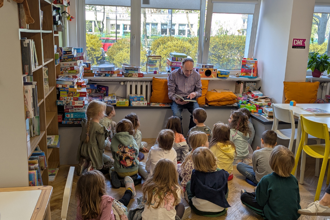 zdjęcie mężczyzny siedzącego i czytającego książkę siedzącym na podłodze dzieciom we wnętrzu biblioteki dla dzieci nr xv. w tle regały biblioteczne.