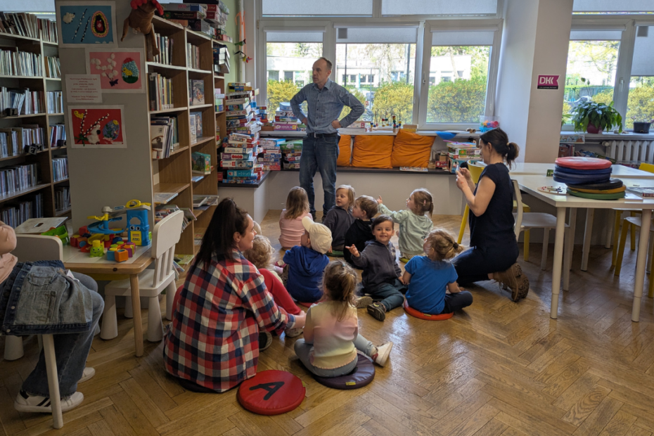 zdjęcie mężczyzny stojącego i czytającego książkę siedzącym na podłodze dzieciom we wnętrzu biblioteki dla dzieci nr xv. w tle regały biblioteczne.