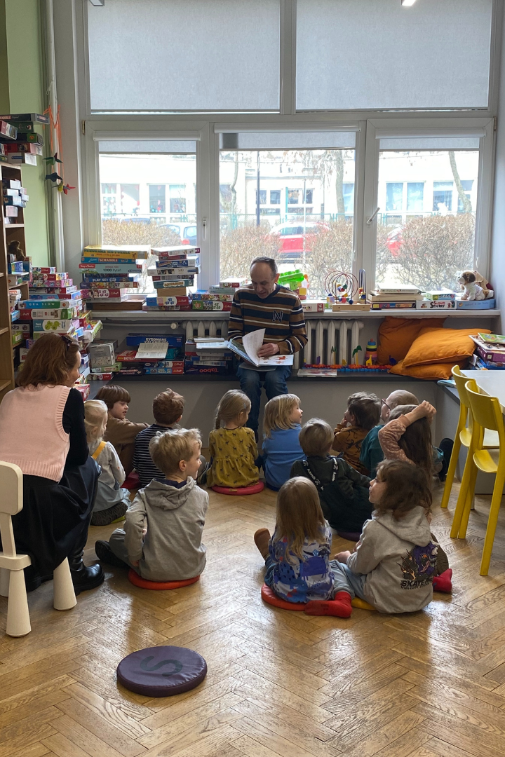 zdjęcie mężczyzny siedzącego i czytającego książkę siedzącym na podłodze dzieciom we wnętrzu biblioteki dla dzieci nr xv. w tle regały biblioteczne.
