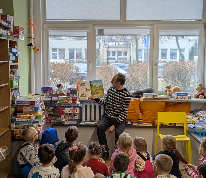 zdjęcie mężczyzny siedzącego i czytającego książkę siedzącym na podłodze dzieciom we wnętrzu biblioteki dla dzieci nr xv. w tle regały biblioteczne.