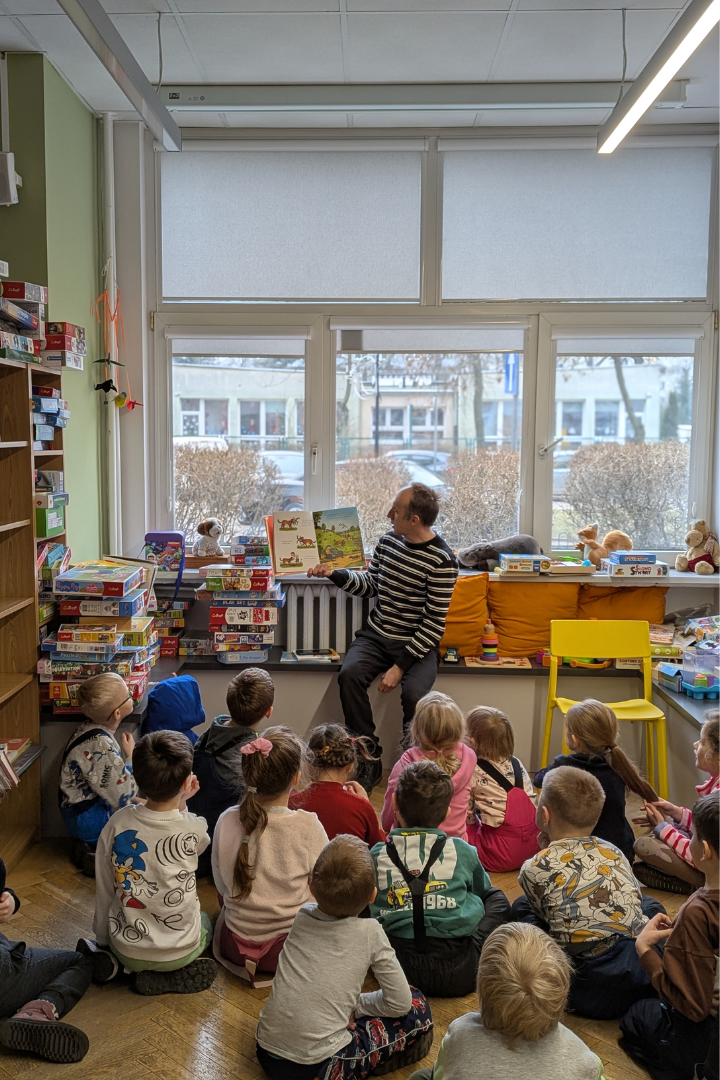 zdjęcie mężczyzny siedzącego i czytającego książkę siedzącym na podłodze dzieciom we wnętrzu biblioteki dla dzieci nr xv. w tle regały biblioteczne.