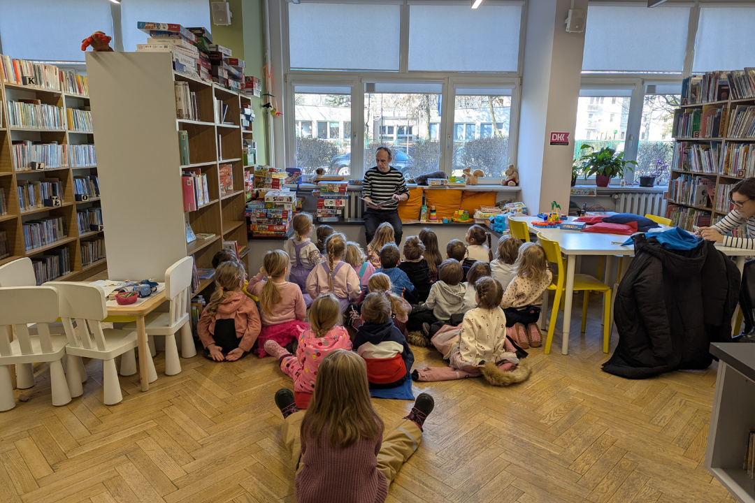 zdjęcie mężczyzny siedzącego i czytającego książkę siedzącym na podłodze dzieciom we wnętrzu biblioteki dla dzieci nr xv. w tle regały biblioteczne.