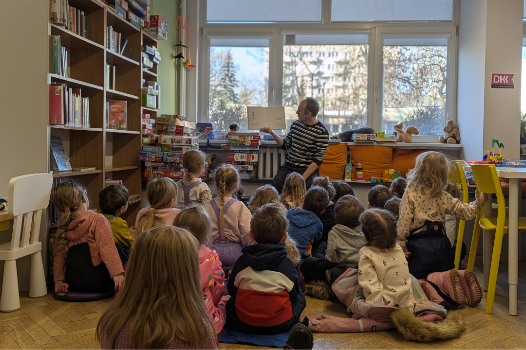 zdjęcie mężczyzny siedzącego i czytającego książkę siedzącym na podłodze dzieciom we wnętrzu biblioteki dla dzieci nr xv. w tle regały biblioteczne.