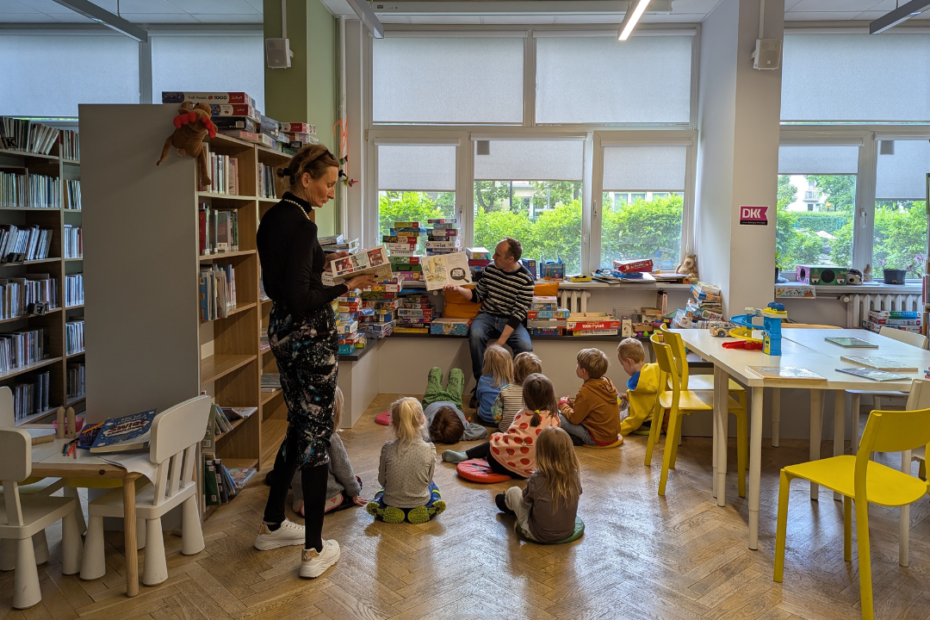 zdjęcie mężczyzny siedzącego i czytającego książkę siedzącym na podłodze dzieciom we wnętrzu biblioteki dla dzieci nr xv. w tle regały biblioteczne.