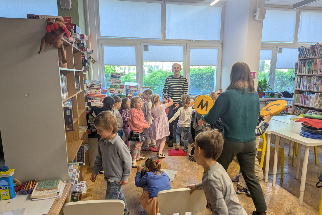 zdjęcie mężczyzny siedzącego i czytającego książkę siedzącym na podłodze dzieciom we wnętrzu biblioteki dla dzieci nr xv. w tle regały biblioteczne.
