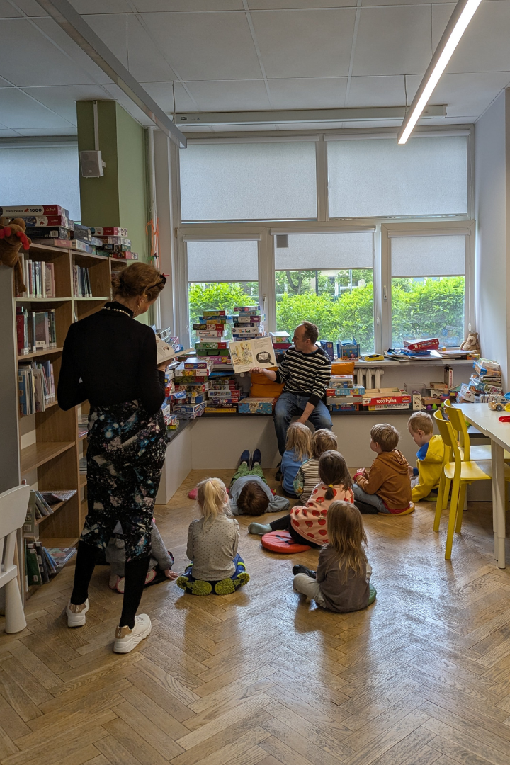 zdjęcie mężczyzny siedzącego i czytającego książkę siedzącym na podłodze dzieciom we wnętrzu biblioteki dla dzieci nr xv. w tle regały biblioteczne.