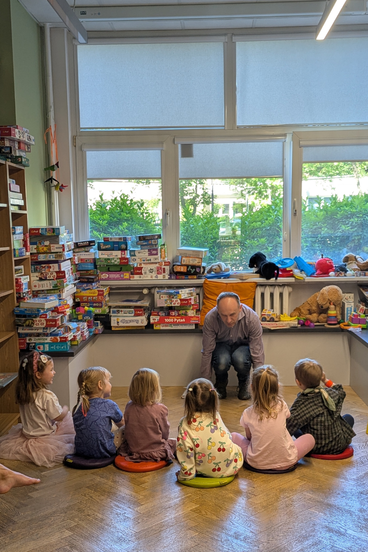 zdjęcie mężczyzny siedzącego i czytającego książkę siedzącym na podłodze dzieciom we wnętrzu biblioteki dla dzieci nr xv. w tle regały biblioteczne.