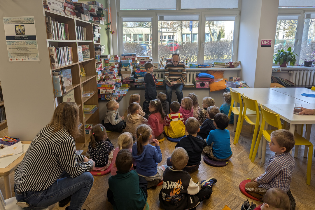zdjęcie mężczyzny siedzącego i czytającego książkę siedzącym na podłodze dzieciom we wnętrzu biblioteki dla dzieci nr xv. w tle regały biblioteczne.