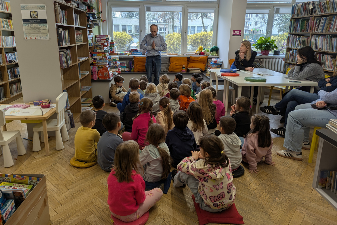 zdjęcie mężczyzny siedzącego i czytającego książkę siedzącym na podłodze dzieciom we wnętrzu biblioteki dla dzieci nr xv. w tle regały biblioteczne.