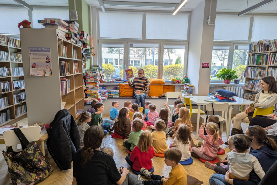 zdjęcie mężczyzny siedzącego i czytającego książkę siedzącym na podłodze dzieciom we wnętrzu biblioteki dla dzieci nr xv. w tle regały biblioteczne.