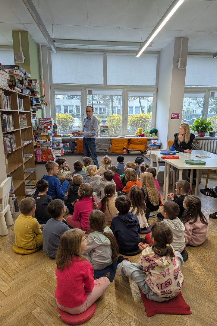 zdjęcie mężczyzny siedzącego i czytającego książkę siedzącym na podłodze dzieciom we wnętrzu biblioteki dla dzieci nr xv. w tle regały biblioteczne.