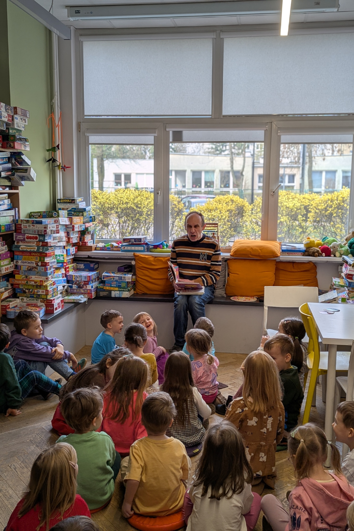 zdjęcie mężczyzny siedzącego i czytającego książkę siedzącym na podłodze dzieciom we wnętrzu biblioteki dla dzieci nr xv. w tle regały biblioteczne.