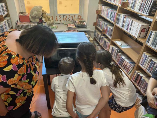 grafika przedstawia dzieci oraz bibliotekarkę. Przyglądają się procesowi tworzenia breloczków, które są zrobione z folii termokurczliwej. Breloczki znajdują się w piekarniku.