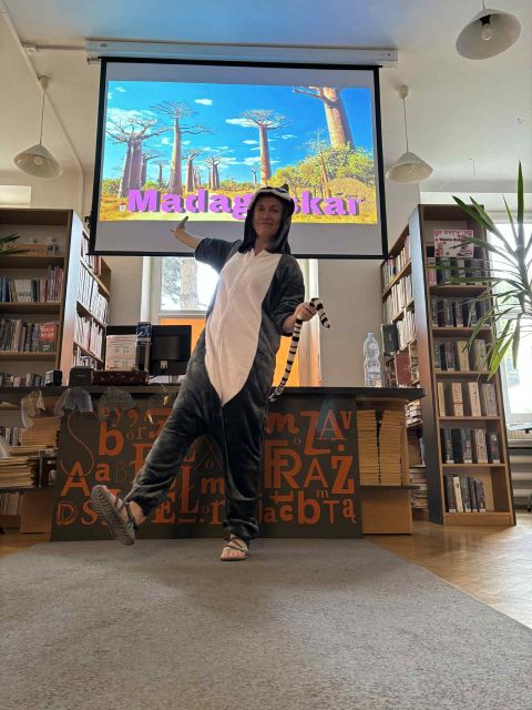 grafika przedstawia bibliotekarkę przebraną w strój lemura, która stoi na tle ekranu. Na Ekranie wyświetlony jest spoczątkowy slajd z baobabami i napisem Madagaskar.