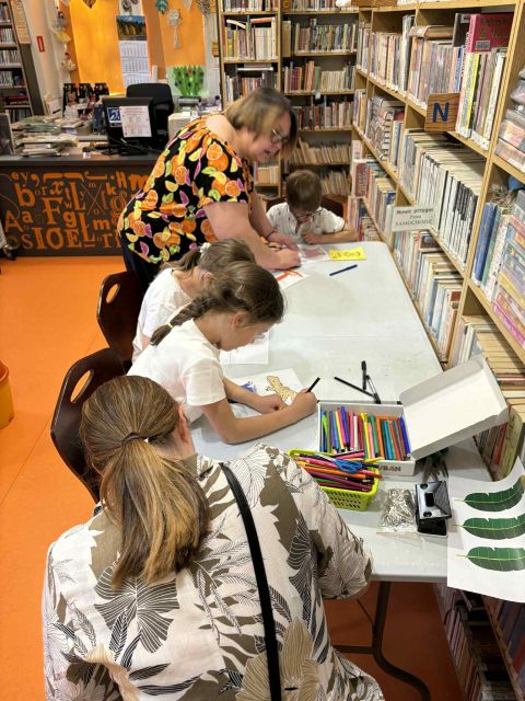 grafika przedstawia dzieci oraz bibliotekarkę w czasie warsztatów plastycznych, na których są wykonywane breloczki z wizerunkami madagaskarskich zwierząt.