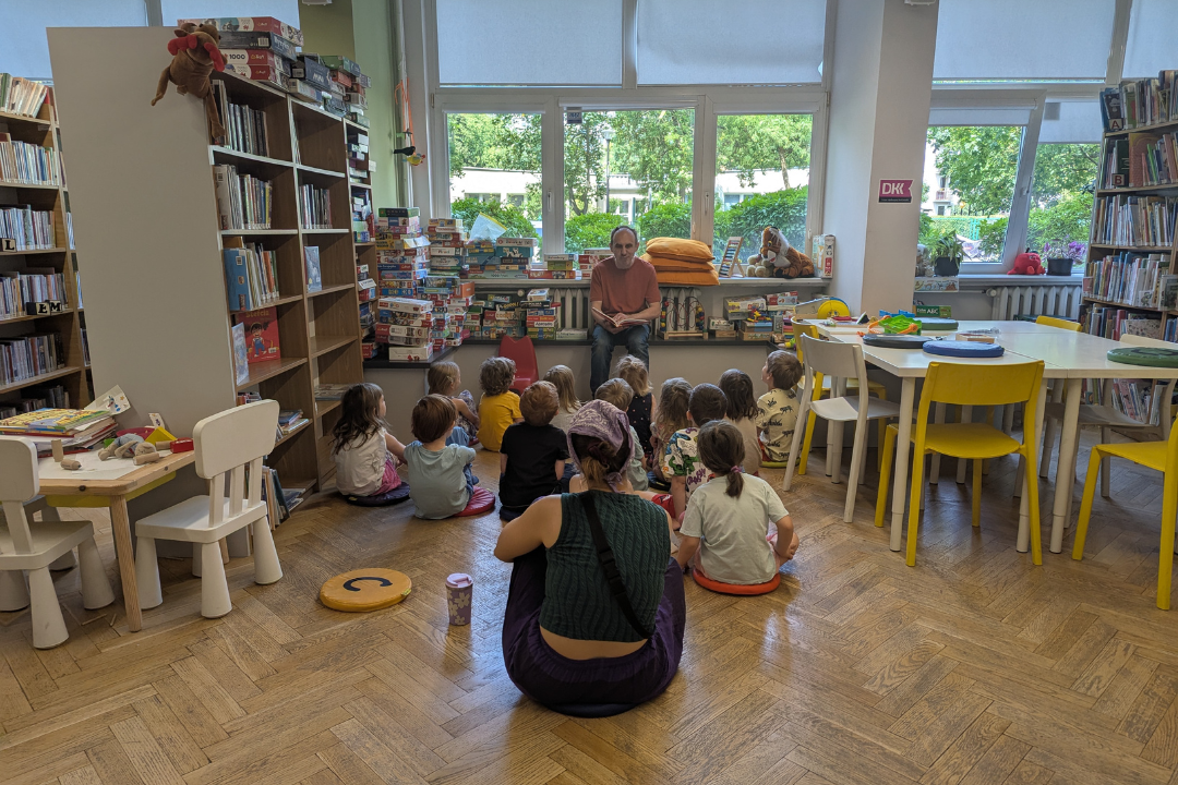 zdjęcie mężczyzny siedzącego i czytającego książkę siedzącym na podłodze dzieciom we wnętrzu biblioteki dla dzieci nr xv. w tle regały biblioteczne.
