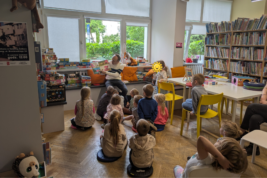 zdjęcie mężczyzny siedzącego i czytającego książkę siedzącym na podłodze dzieciom we wnętrzu biblioteki dla dzieci nr xv. w tle regały biblioteczne.