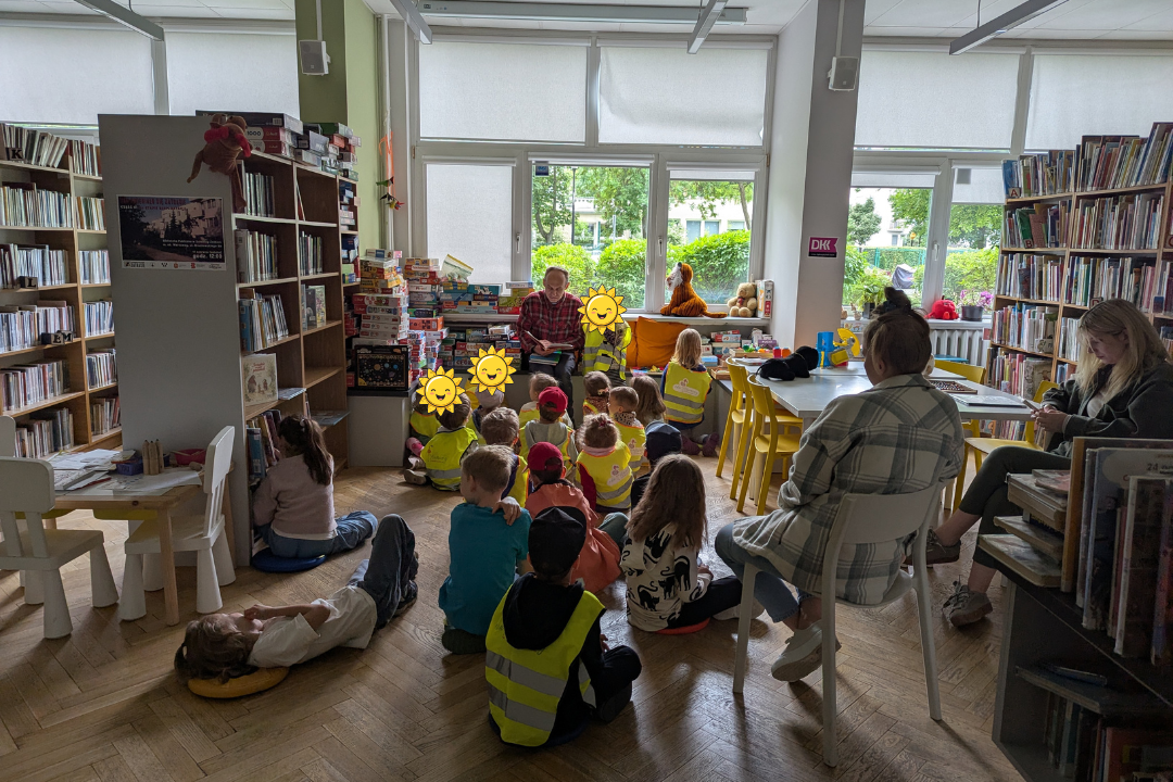 zdjęcie mężczyzny siedzącego i czytającego książkę siedzącym na podłodze dzieciom we wnętrzu biblioteki dla dzieci nr xv. w tle regały biblioteczne.