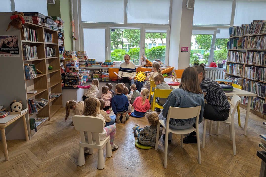 zdjęcie mężczyzny siedzącego i czytającego książkę siedzącym na podłodze dzieciom we wnętrzu biblioteki dla dzieci nr xv. w tle regały biblioteczne.