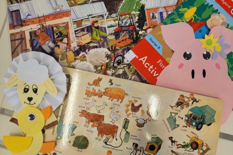 Na białym stole książki z obrazkami dla dzieci (farma, wieś). Obok różowa świnka, biała owca i żółta kaczka z papieru.