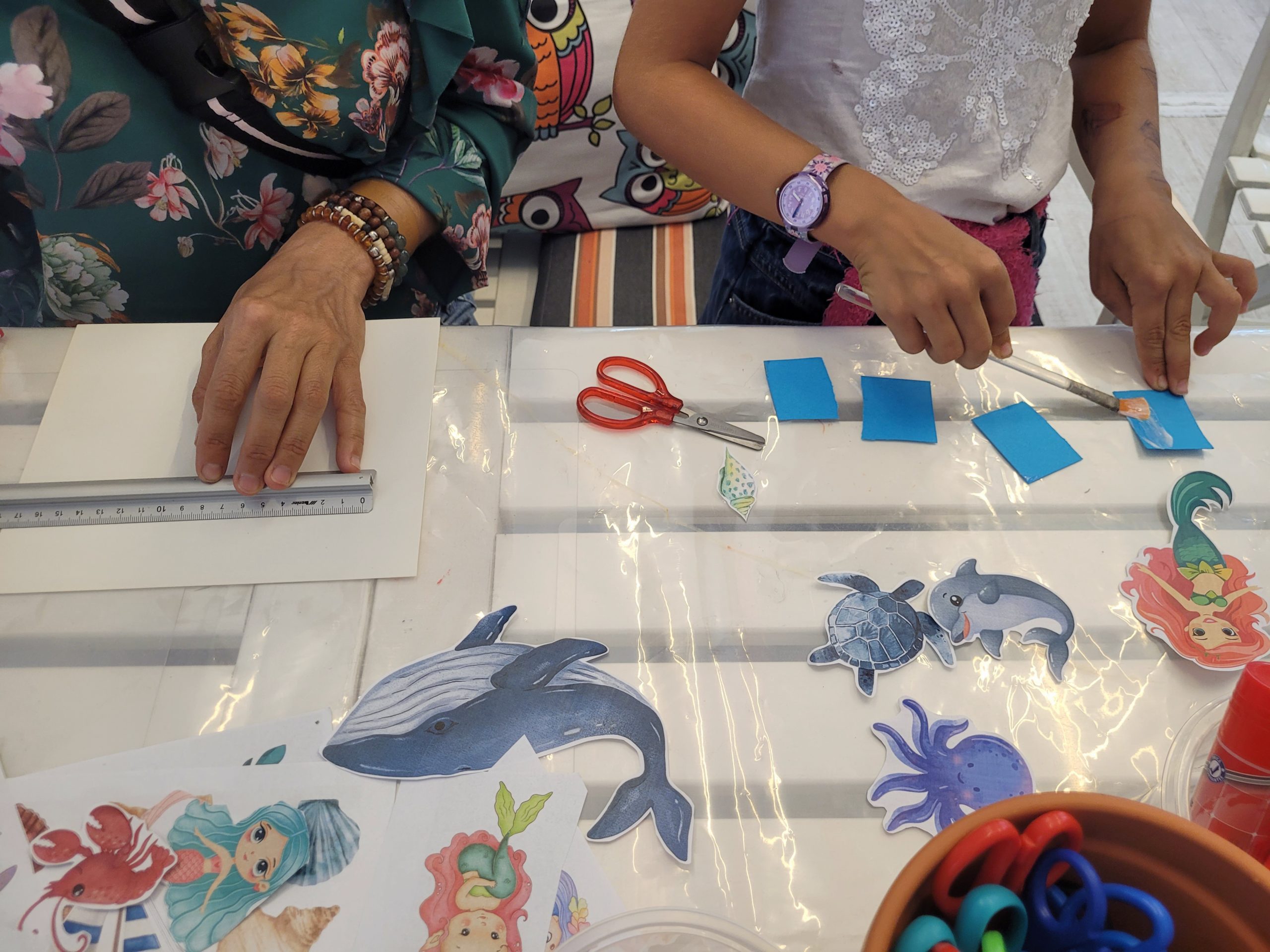 Przy stole kobieta i dziecko (twarzy nie widać) wykonują prace plastyczne. Na stole papier, nożyczki, naklejki z tematem morza.