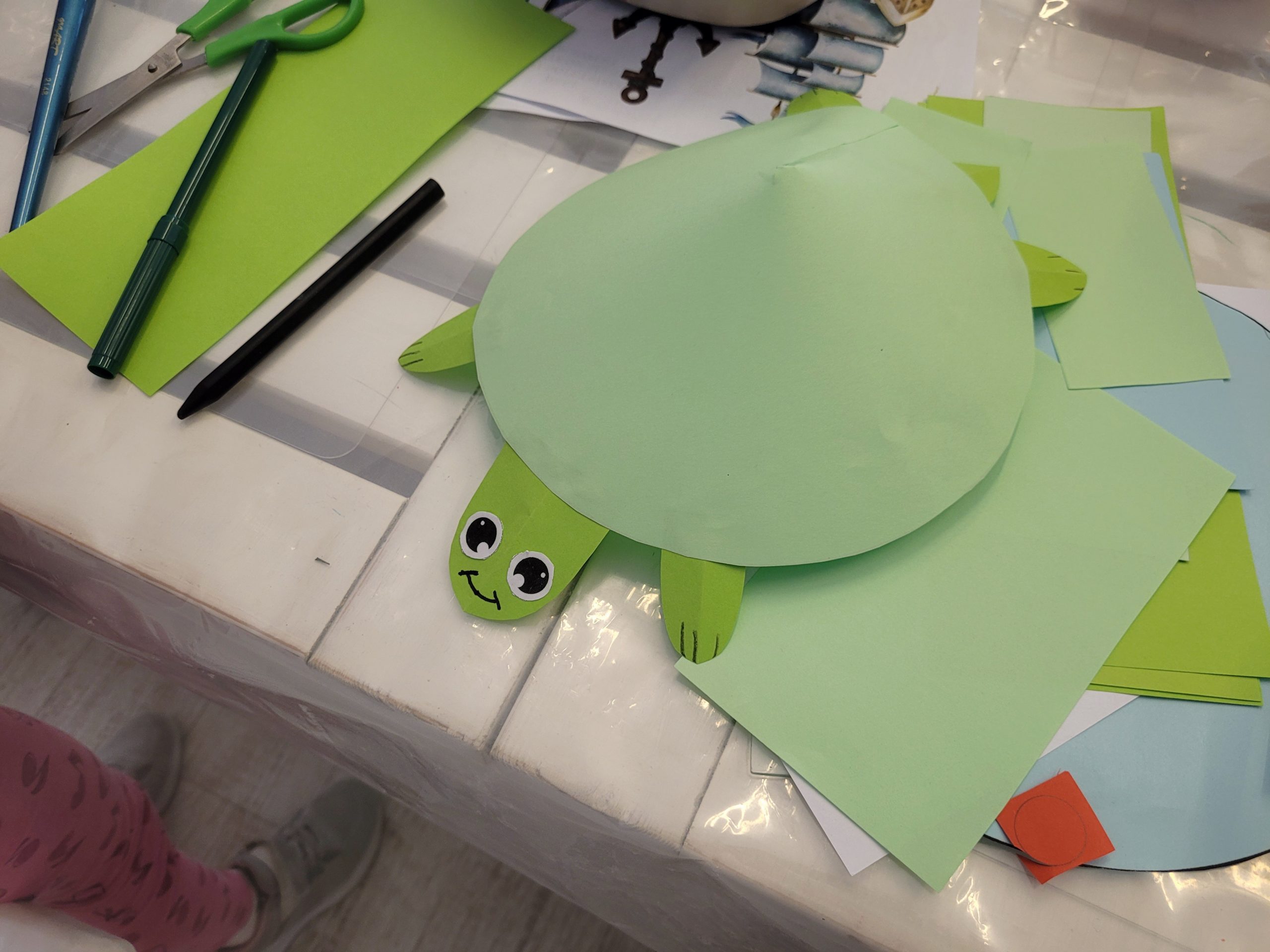 Na stole zielony papierowy żółw, nożyczki, flamastry i zielony papier. 