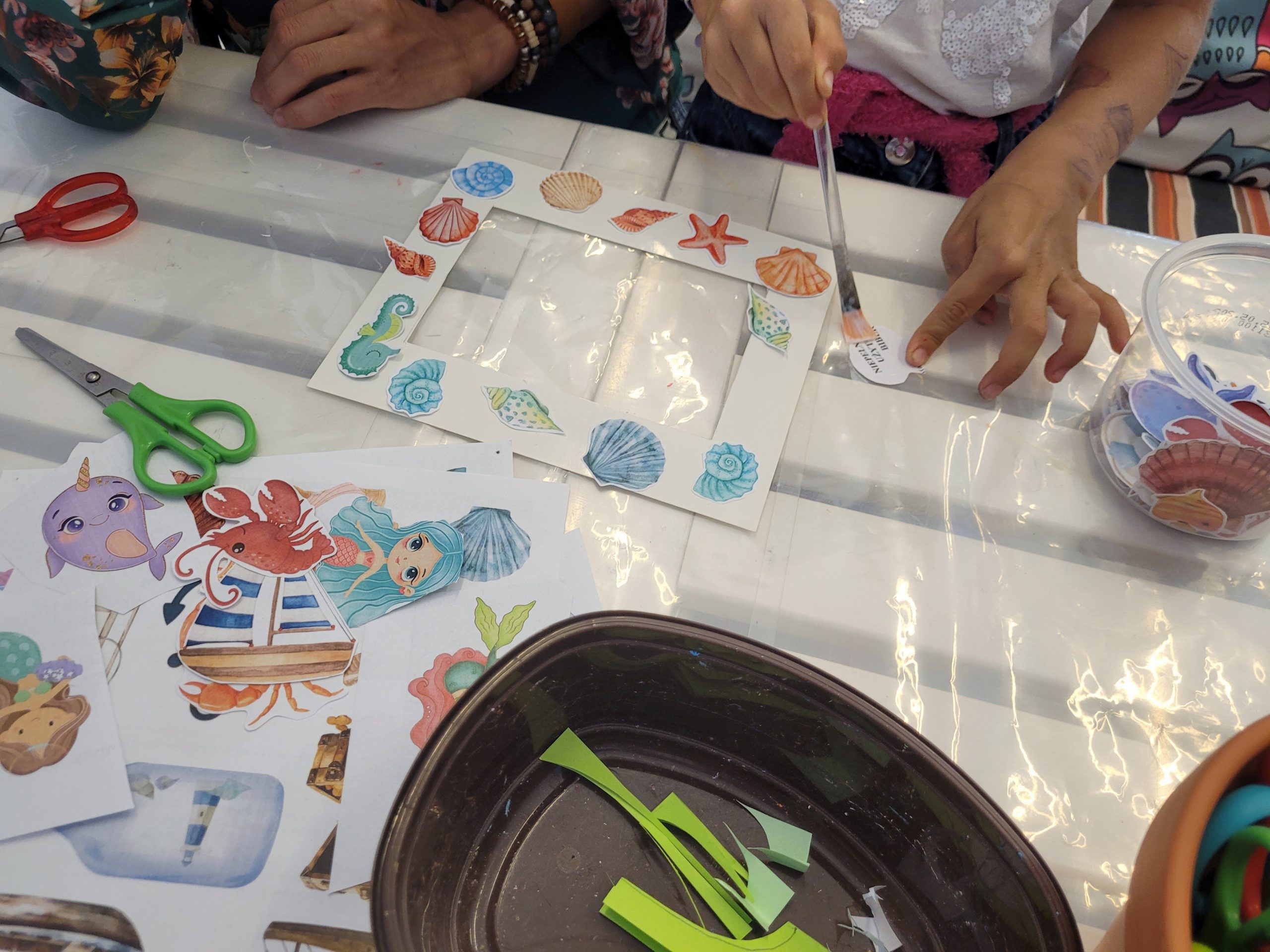 Przy stole kobieta i dziecko wykonują papierową ramkę z muszelkami. Na stole kolorowe naklejki z tematem morza, nożyczki, klej.