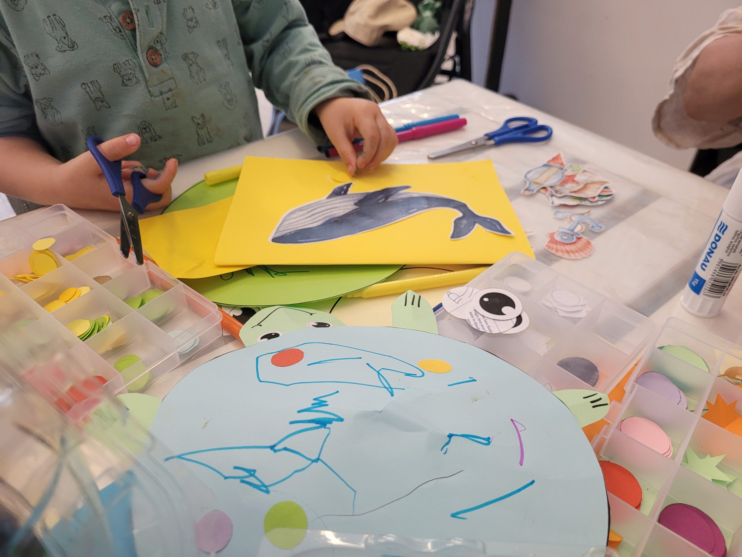 Przy stole dzieci wykonują papierowe prace. Dziecko z nożyczkami robi żółtą kartkę z wielorybem. Na stole papier i pudełko z kolorowymi kółeczkami.