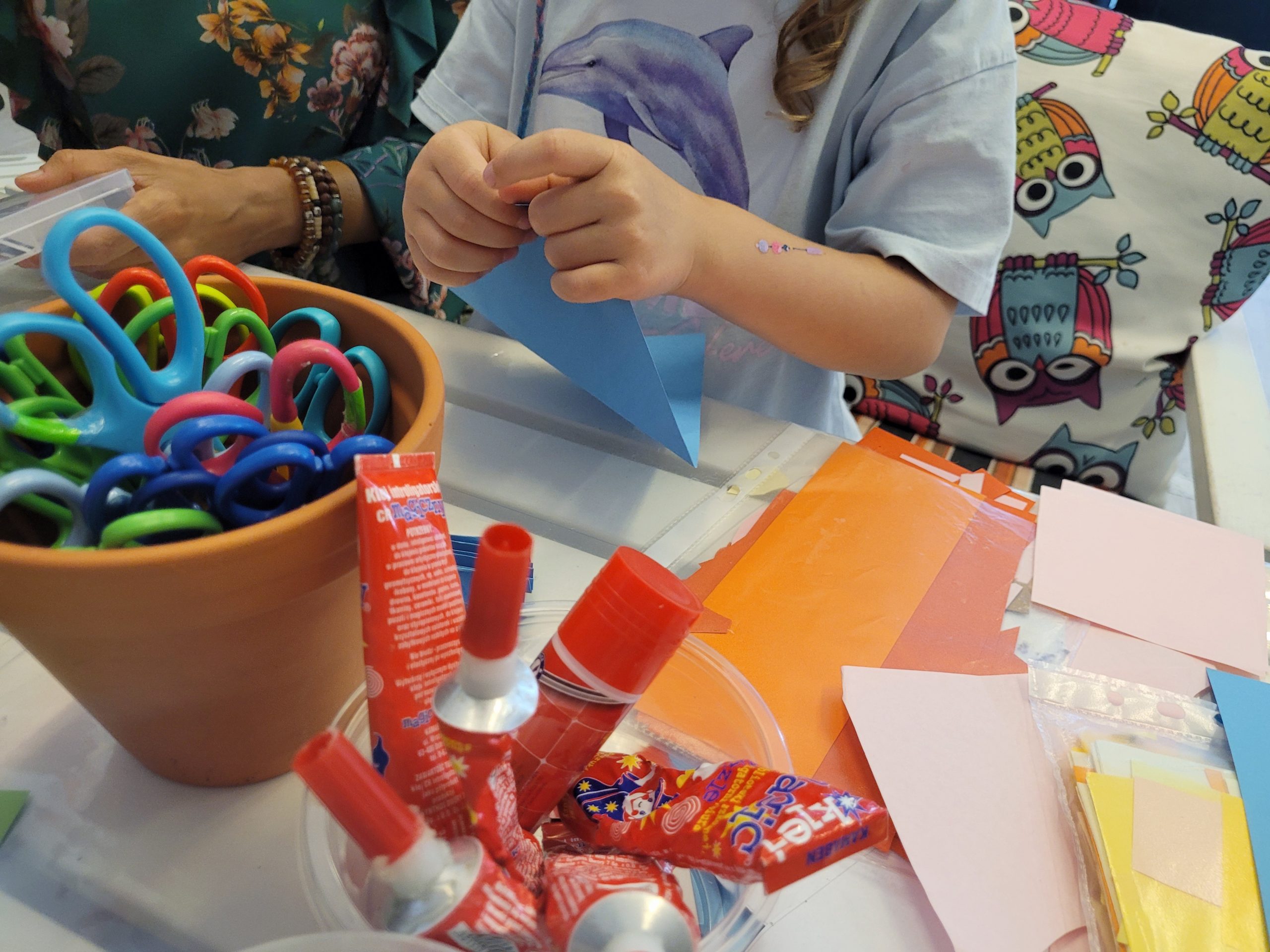 Dziewczynka i kobieta (widać tylko ręce) wykonują prace z papieru. Na stole nożyczki, kleje, kolorowy papier.