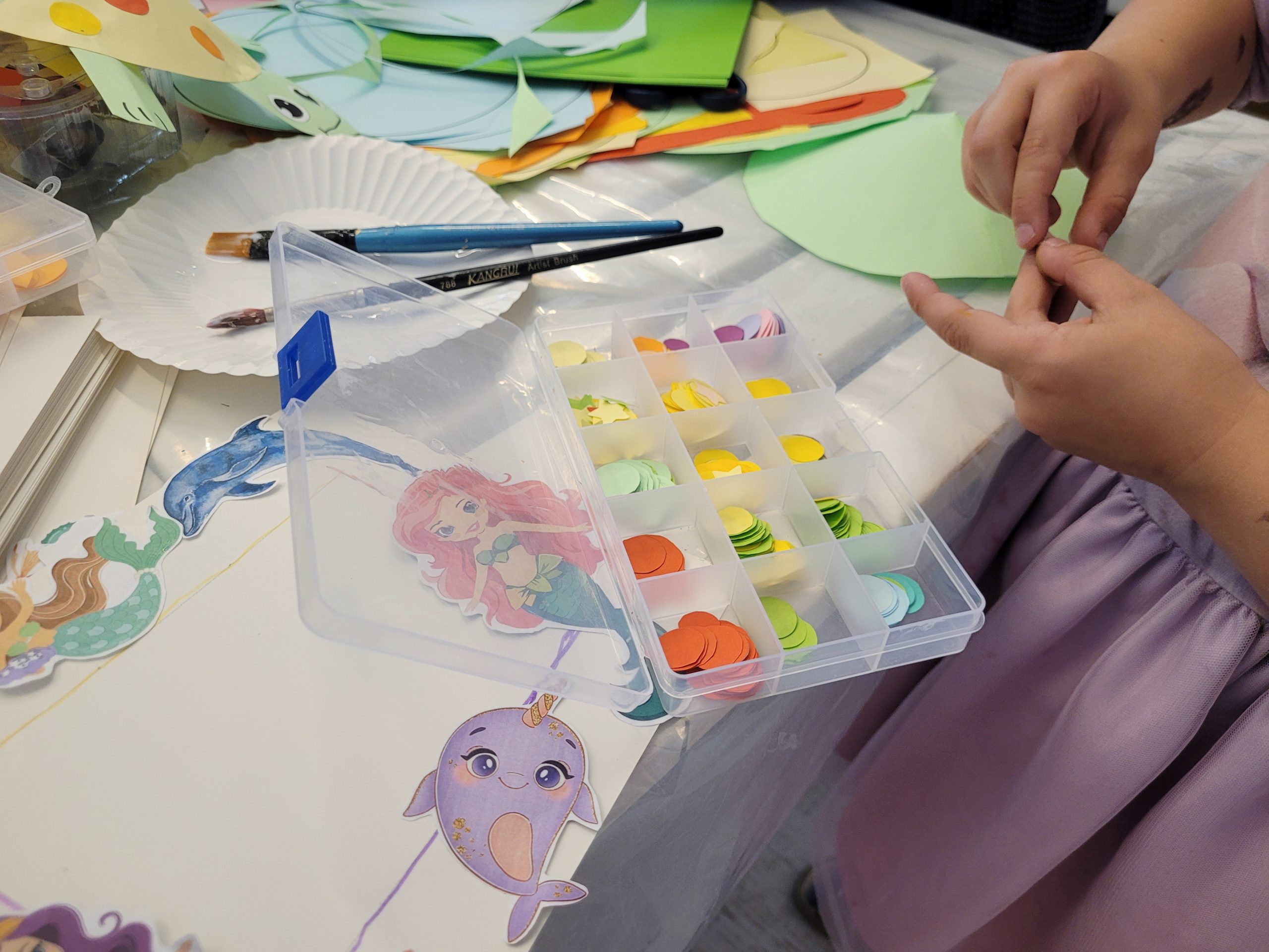 Dziecko (twarzy nie widać) wykonuje pracę plastyczne. Na stole kolorowy papier, miseczka z klejem, naklejki morskie, pudełko z kolorowymi kółkami.