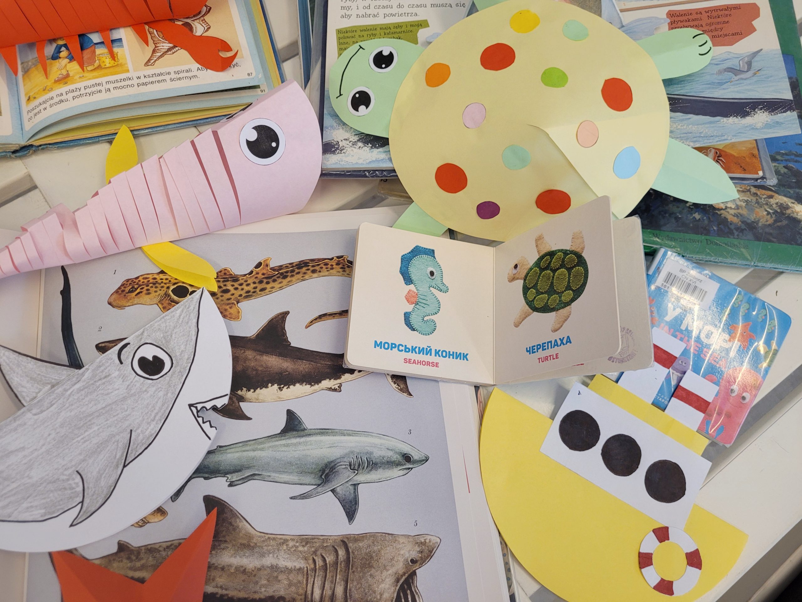 Na stole kolorowe książki dla dzieci z tematem morza oraz papierowe dzieła: rekin, różowa ryba, żółto-zielony żółw, żółty statek.