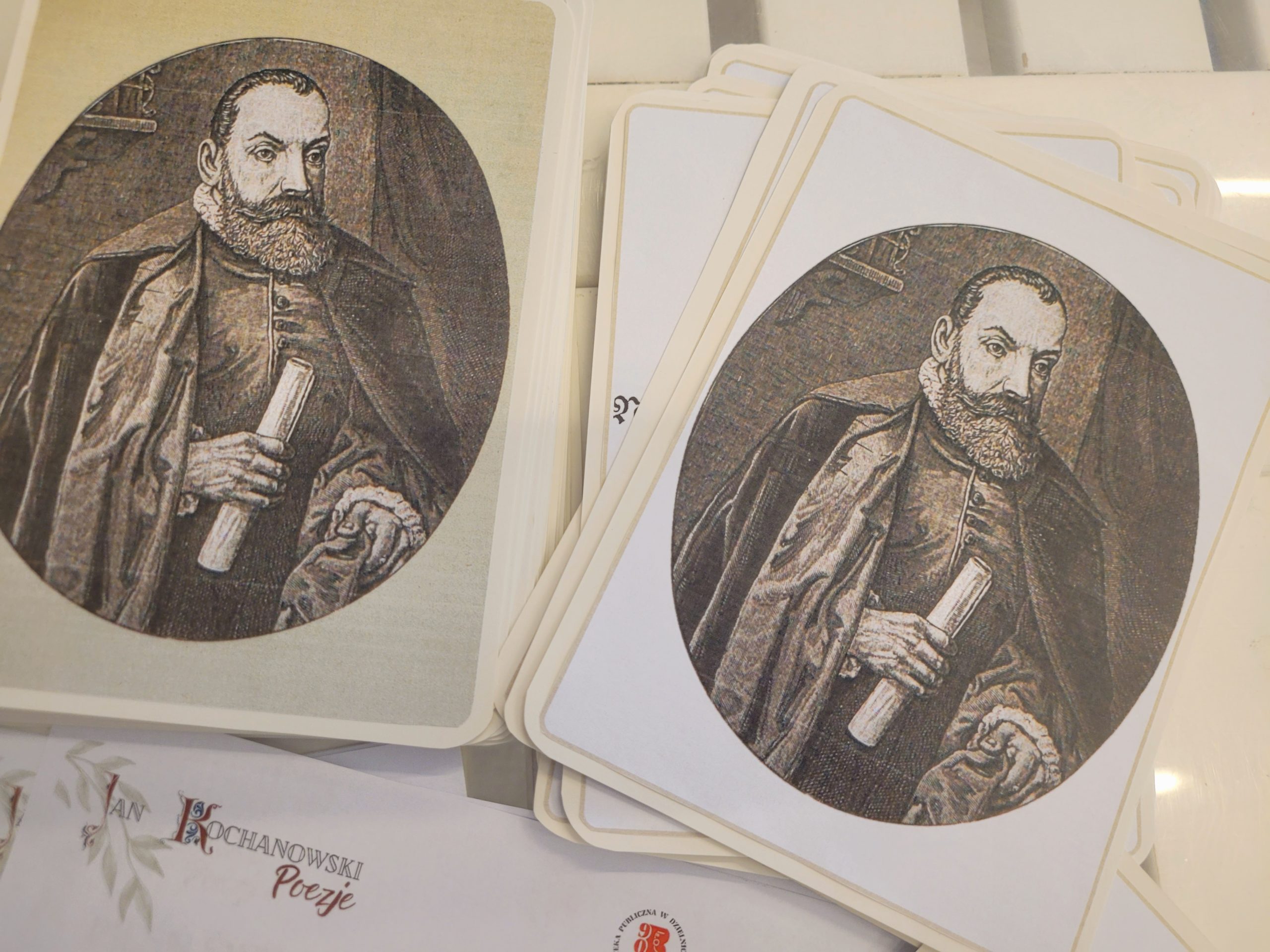 Na stole karty przedstawiające grafikę mężczyzny w stroju renesansowym, niżej kartka, w rogu której tekst Jan Kochanowski Poezje