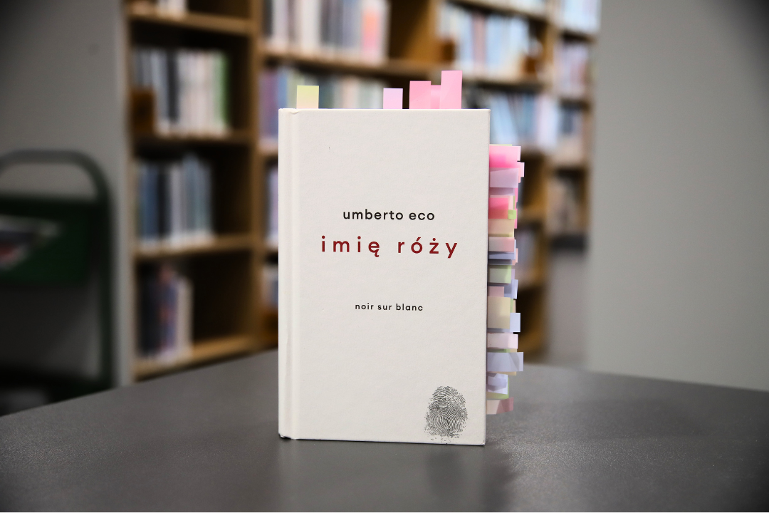 zdjecie przedstawia okładkę książki imię rózy umberto eco na tle wnętrza wypożyczalni nr 138, widoczne napisy: umberto eco imię róży noir sur blanc